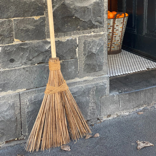 Strohbesen neben einem Hauseingang, angelehnt an die Fassade aus Steinen. Im Hauseingang stehend ein grosser Korb mit Orangen oder Mandarinen.